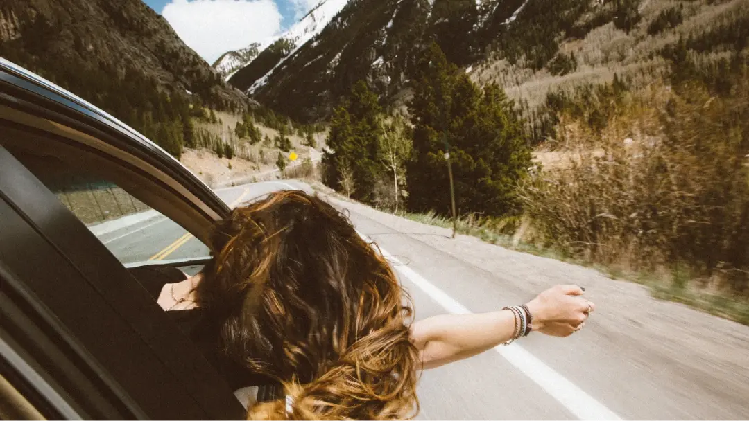 Una mujer disfruta de la brisa sacando el brazo y la cabeza por la ventanilla delantera-derecha de un cohe en marcha mientras al fondo se observa un paisaje montañoso.